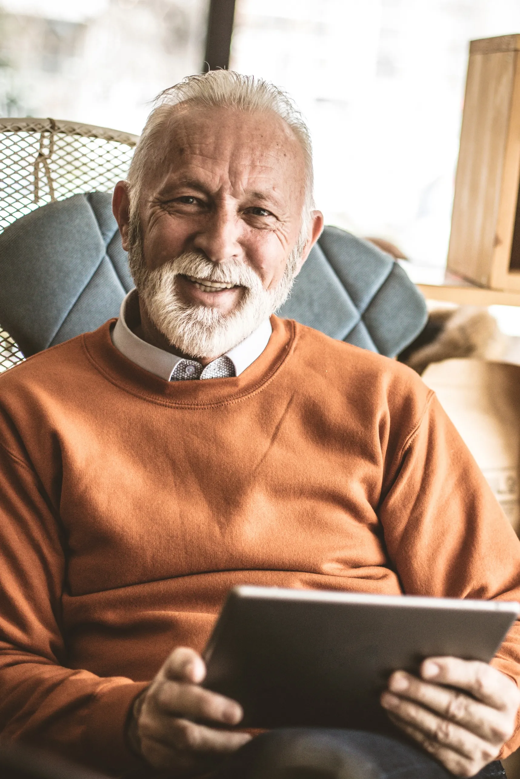 Älterer Herr im orangefarbenen Pullover sitzt lächelnd mit einem Tablet in den Händen.