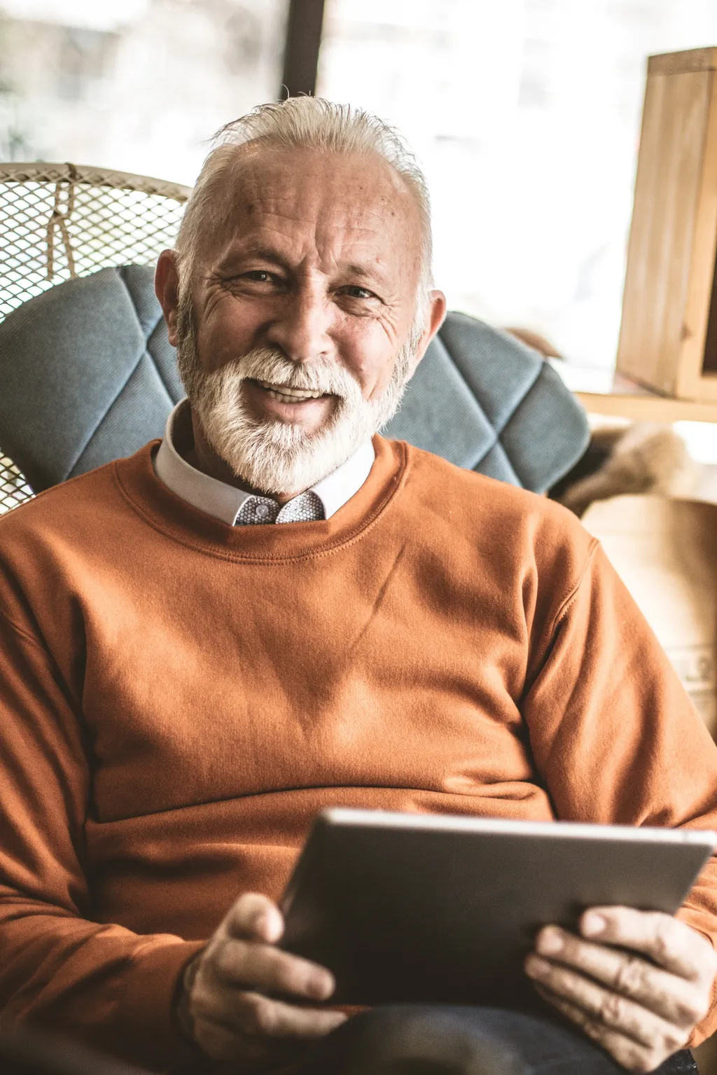 Älterer Herr im orangefarbenen Pullover sitzt lächelnd mit einem Tablet in den Händen