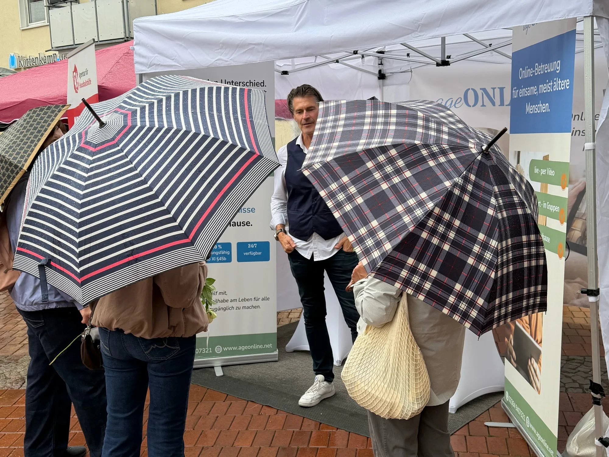 Besucherinnen und Besucher mit Regenschirmen stehen vor dem ageONLINE-Stand auf dem Marktplatz