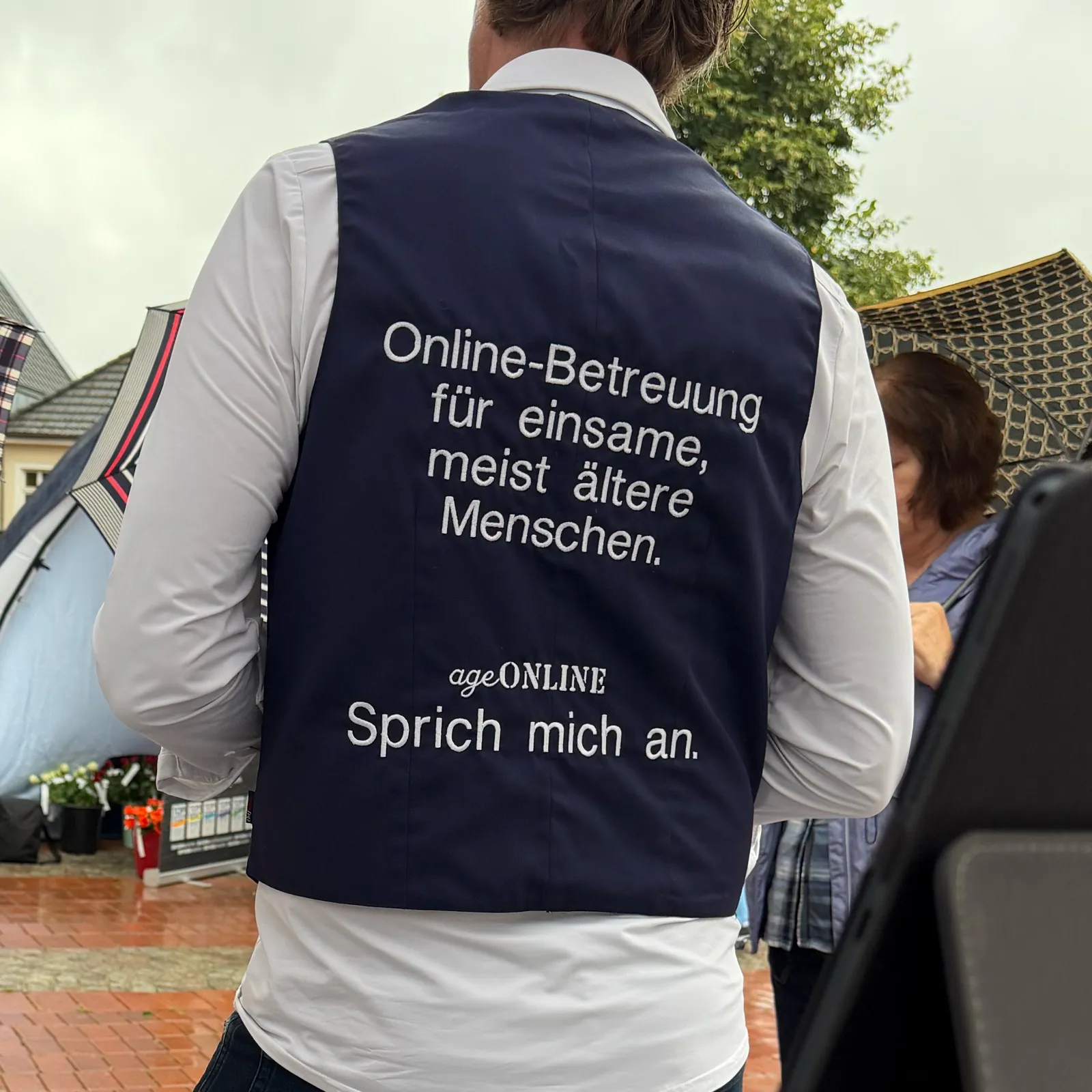 Rückenansicht einer dunkelblauen Weste mit der Aufschrift: Online-Betreuung für einsame, meist ältere Menschen. ageONLINE. Sprich mich an.