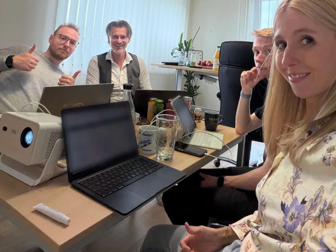 Das Team sitzt mit Laptops am Besprechungstisch und lacht in die Kamera