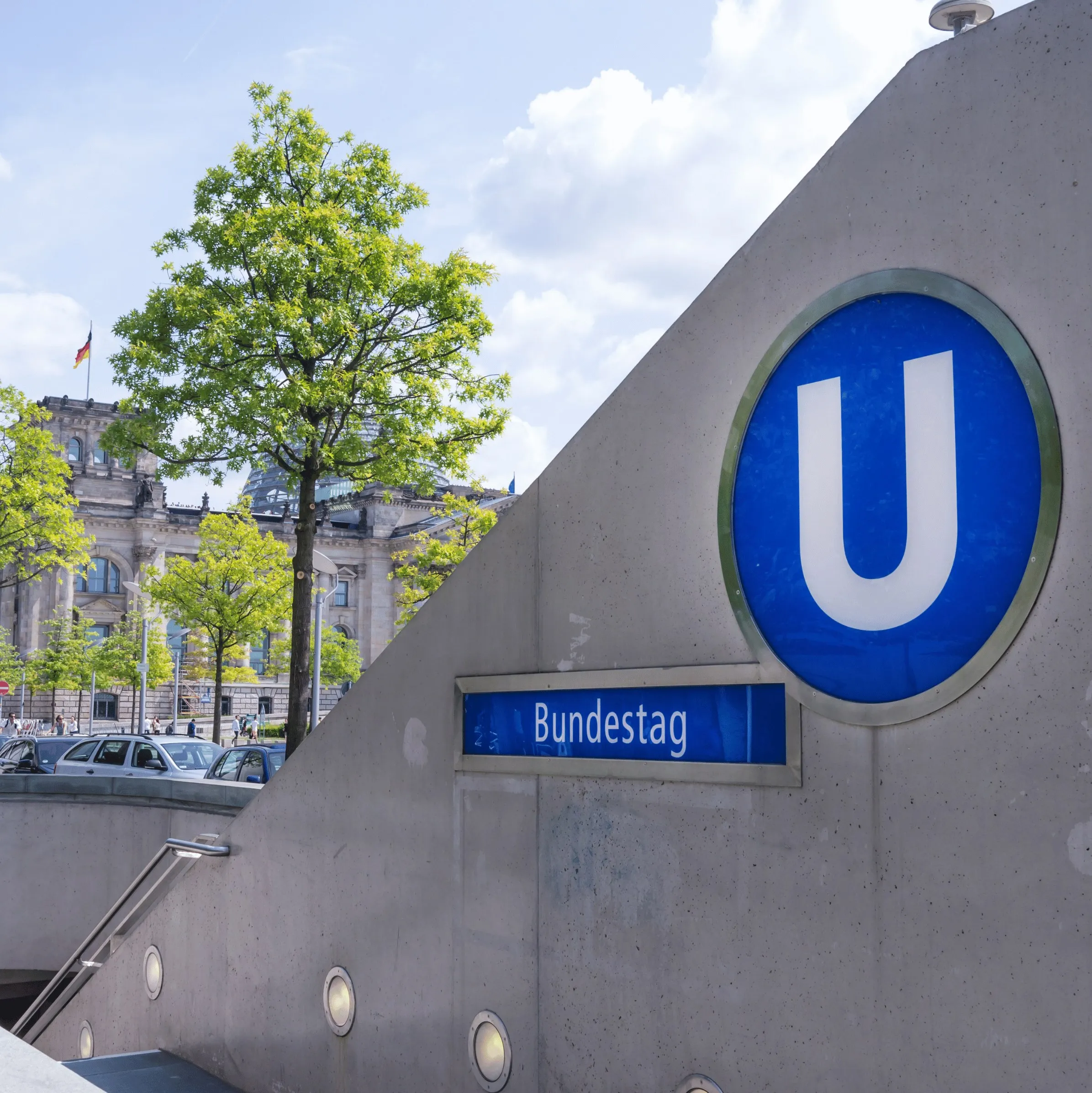 Schild der U-Bahn-Station Bundestag in Berlin, im Hintergrund das Reichstagsgebäude hinter Bäumen.